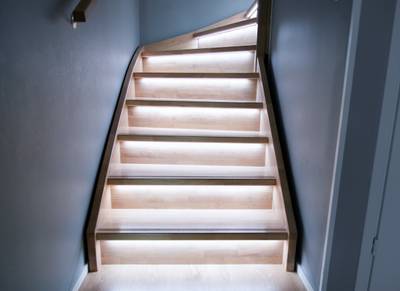 Un escalier moderne en bois avec des lumières LED sous chaque marche, mettant en valeur le design minimaliste avec un éclairage chaleureux sur des murs gris.