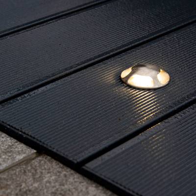 Lampe de terrasse ronde de couleur argentée avec des planches de bois rainurées foncées, émettant une douce lueur la nuit. Idéal pour les solutions d'éclairage LED extérieures.