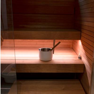 Intérieur de sauna en bois avec un seau et une pelle en métal sur les bancs ; un éclairage LED chaleureux met en évidence les raisons de la présence du bois sur les surfaces d'assise.