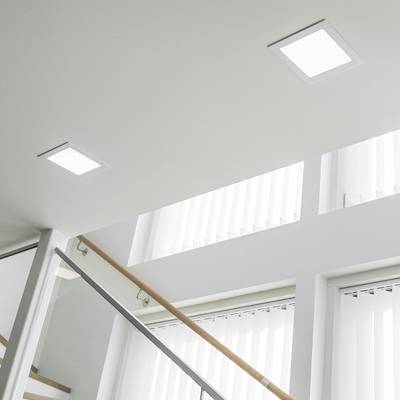 Des plafonniers encastrés carrés lumineux éclairent un escalier moderne avec une balustrade en verre et de grandes fenêtres avec des rideaux verticaux.