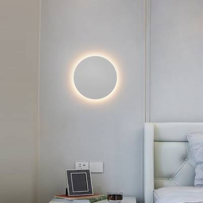 Une applique circulaire moderne illumine une chambre à coucher minimaliste avec des murs gris clair, une tête de lit touffetée blanche et une décoration de table de nuit élégante. Boutique d'éclairage LED.