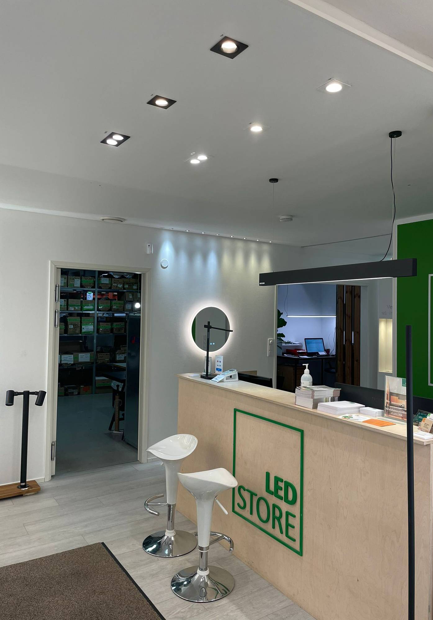 Luminaires à Led dans la salle d\'exposition de Ledstore. Des spots au plafond devant le comptoir et un plafonnier VIIVA au-dessus du comptoir. Le mur est accentué par des spots de 3W, fixes et directionnels. Miroir lumineux Moon 600 sous les spots. Ledstore.fi