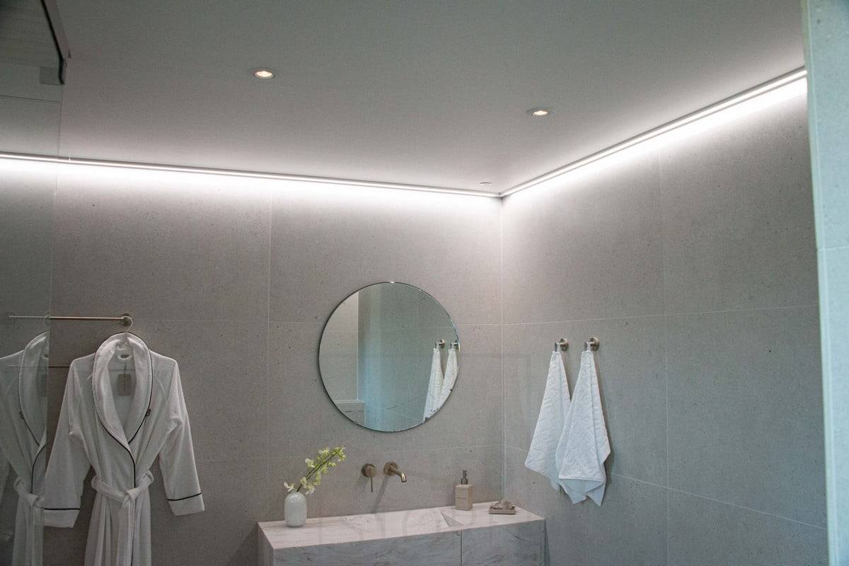 Housing Fair 2022 : Bande LED montée sur une surface basse, au plafond près du mur, fournissant une lumière douce et indirecte à la salle de bains. Ledstore.fi
