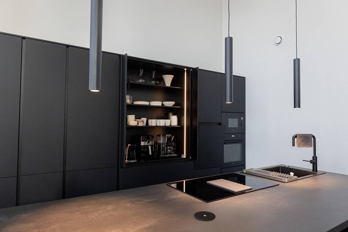 Dans la cuisine, trois suspensions noires PIIPPU 2.0 sont suspendues au-dessus de l'îlot. De plus, à l'arrière, le placard à petit-déjeuner est éclairé par une bande LED installée à la verticale.