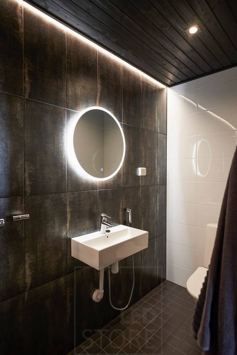 Salle de bain sombre éclairée de façon atmosphérique par des spots et de la lumière indirecte. Le miroir lumineux est doté d'une fonction de désembuage qui permet de conserver la luminosité du miroir en sortant du sauna ou de la douche. Ledstore.fi