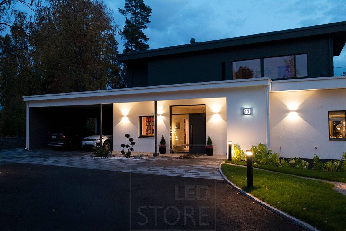 Applique blanche bidirectionnelle FUNK éclairant la façade et l'entrée. L'allée est éclairée par des luminaires à colonne. Ledstore.fi
