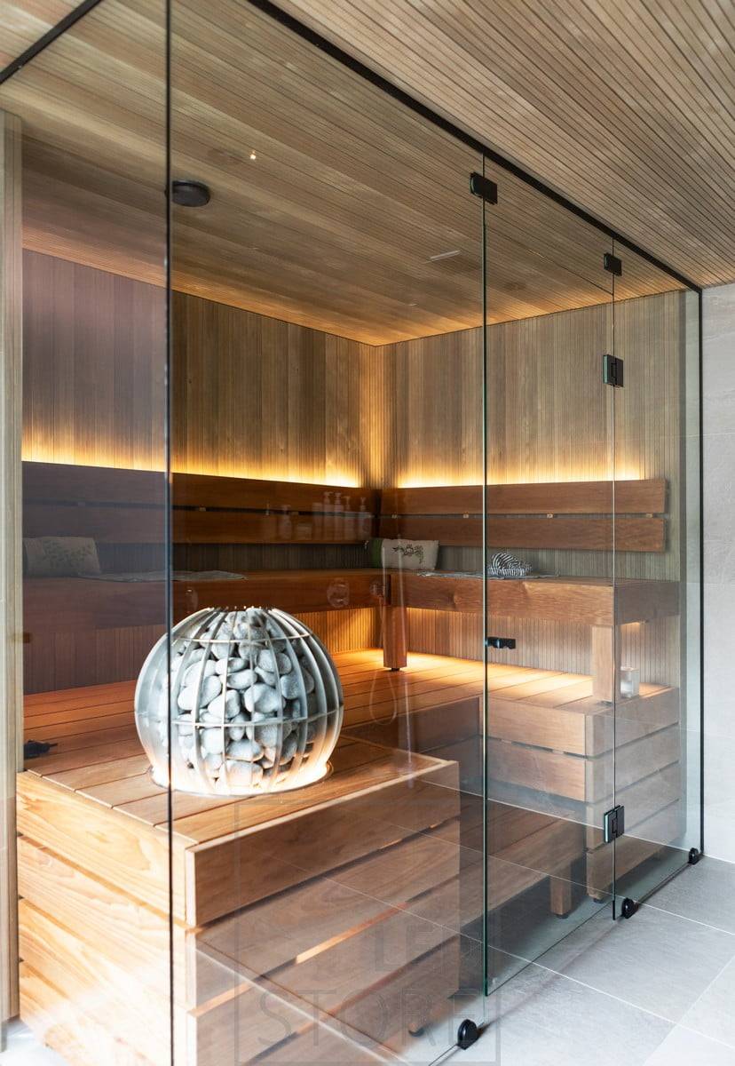 Un sauna aux parois de verre avec une bande lumineuse LED pour créer une lumière indirecte derrière le dossier, sous le plafond et sous le poêle.