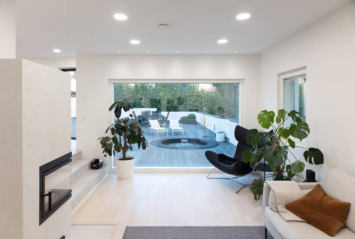 Un salon avec une surface claire et une grande fenêtre donnant sur l'extérieur. Il y a des panneaux LED ronds au plafond et de petits spots devant la fenêtre.