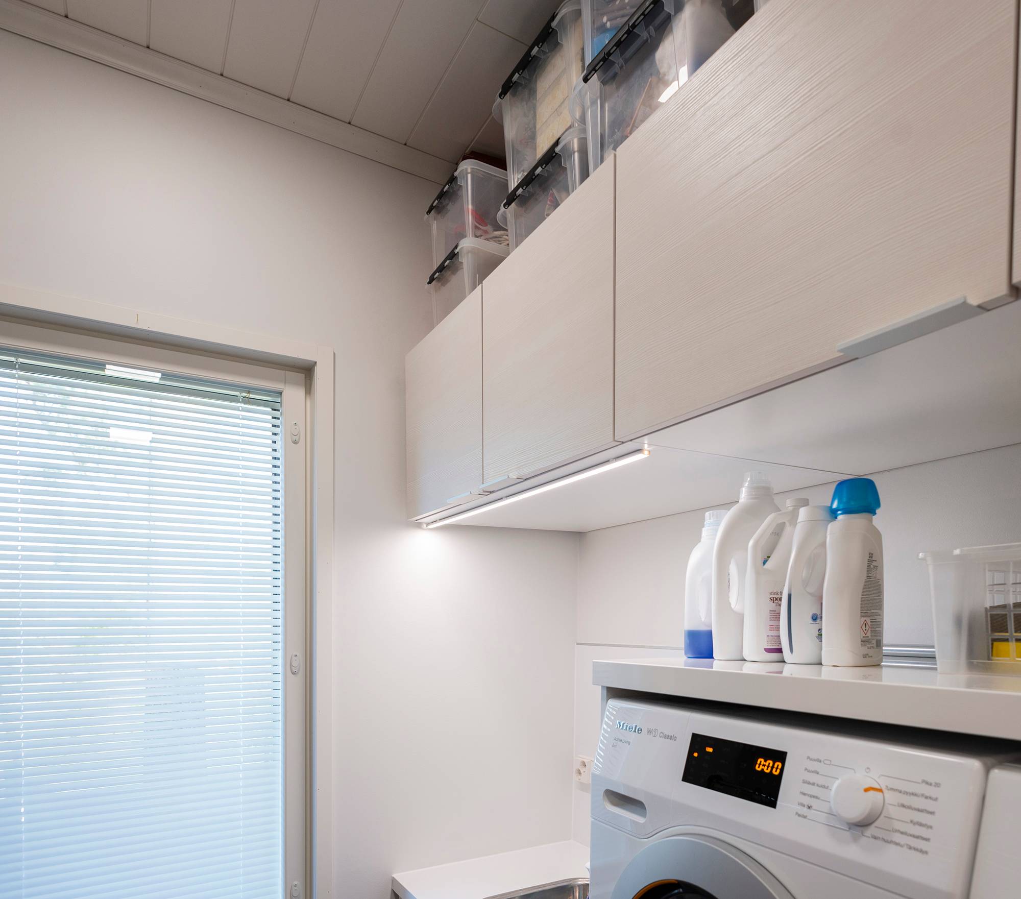 Bande LED pour un éclairage supplémentaire dans une buanderie, fixée sur le bord avant d\'une armoire. Ledstore.fi