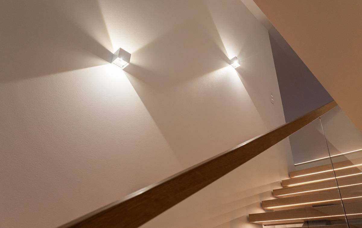 Un escalier moderne avec des marches en bois et une rampe en verre est mis en valeur par les élégantes appliques LED de LedStore.fi, qui créent de magnifiques motifs lumineux sur le mur. Choisissez des luminaires design qui donneront à votre maison un aspect moderne ! 