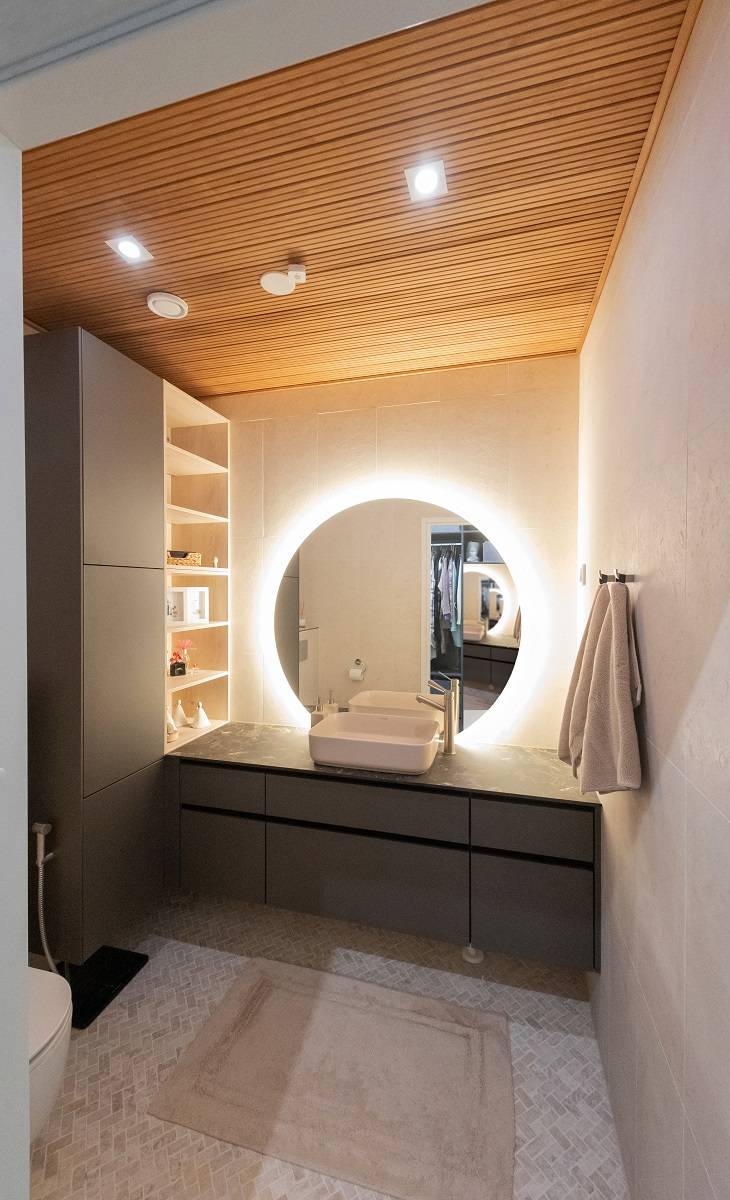 Une salle de bain moderne avec l'éclairage LED de LedStore.fi : un miroir circulaire rétro-éclairé par LED au-dessus du lavabo crée une atmosphère élégante. La salle de bains est composée d'armoires sombres, de plafonds en bois chaleureux et de murs clairs. Les serviettes sont placées sur le côté droit et des étagères vous attendent sur le côté gauche. Le sol est recouvert d'une moquette beige qui ajoute à l'effet d'ensemble.   