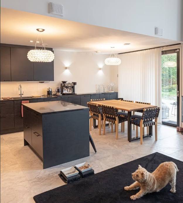 La cuisine et la salle à manger modernes, avec leurs armoires noires, leur table en bois et leur éclairage LED LedStore.fi confortable, créent une atmosphère agréable. Un chat s'étirant sur un tapis noir observe l'ensemble. 