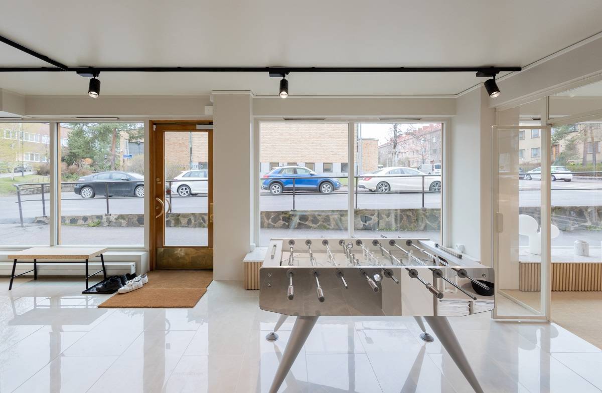 Un éclairage de bureau simple et efficace pour la garantie immobilière Un intérieur moderne avec de grandes fenêtres qui permettent de voir les voitures garées à l'extérieur, avec un baby-foot sur le sol brillant. Un banc avec une étagère à chaussures et un tapis de porte est placé sous un éclairage LED sur rail, soulignant le design moderne de la pièce. Vous trouverez tout cela dans la vaste gamme de solutions d'éclairage LED de LedStore.fi pour votre intérieur.