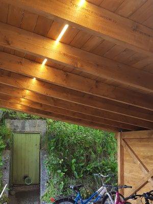 Auvent en bois, éclairé par une bande LED passant entre le toit et les parapets du toit. Des vélos sous l'auvent et une porte verte en arrière-plan. 