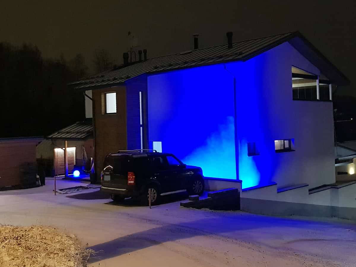 La façade d'une maison moderne éclairée par des LED bleues la nuit. Une voiture dans la cour et une légère couche de neige au sol.