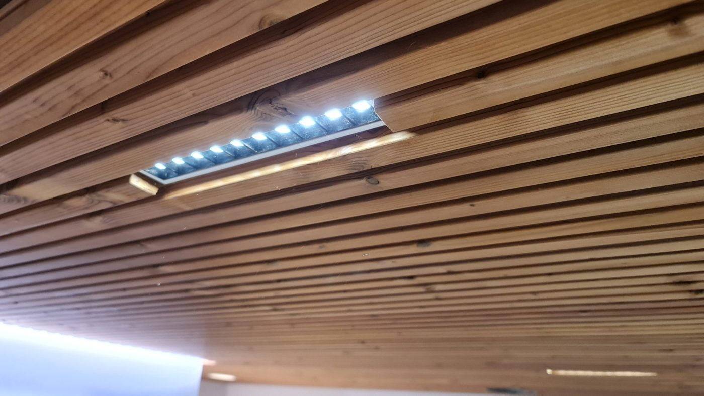 Le plafond en bois avec des lattes parallèles comprend un éclairage LED encastré. Les LED sont partiellement visibles à travers une ouverture rectangulaire, créant une lueur subtile sur la surface en bois naturel.