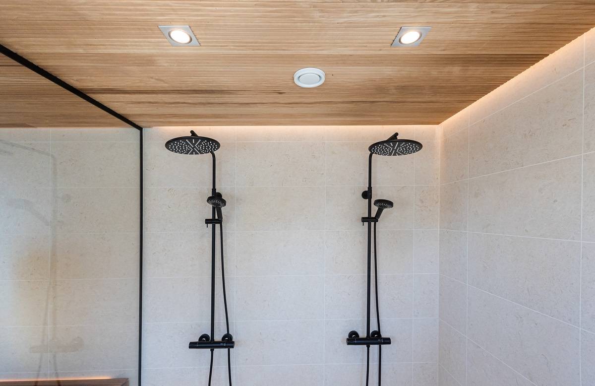 La salle de douche moderne est équipée de deux pommeaux de douche noirs pour un carrelage de couleur crème. Le plafond lambrissé, rehaussé par des lampes LED encastrées de LedStore.fi, crée un look minimaliste et élégant. La douche est séparée du reste de l'espace par une paroi extérieure en verre.  