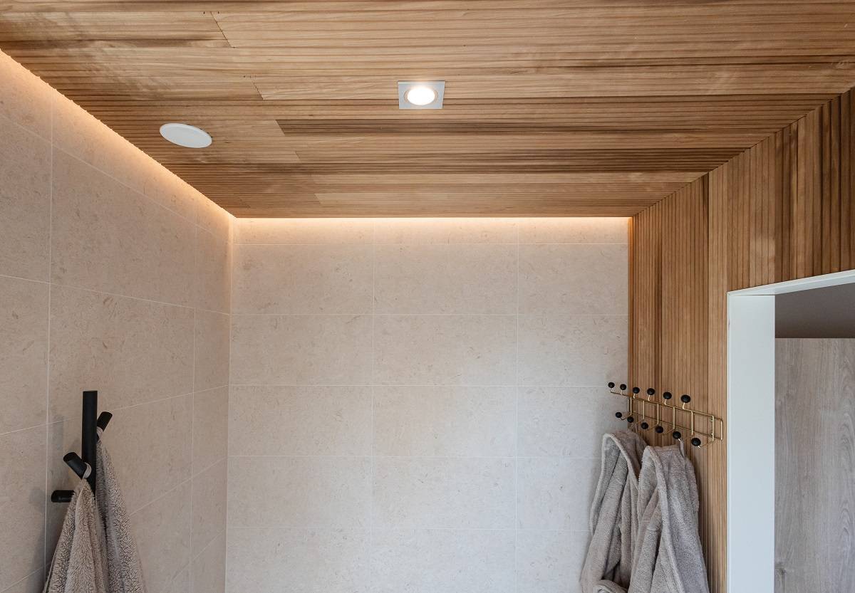 LedStore.fi propose une solution pour une salle de bain moderne avec un éclairage LED moderne. Les murs carrelés beiges, le plafond en bois et l'éclairage LED encastré créent un look frais et moderne. Les serviettes de bain grises et douces suspendues à des crochets noirs harmonisent l'intérieur minimaliste.  