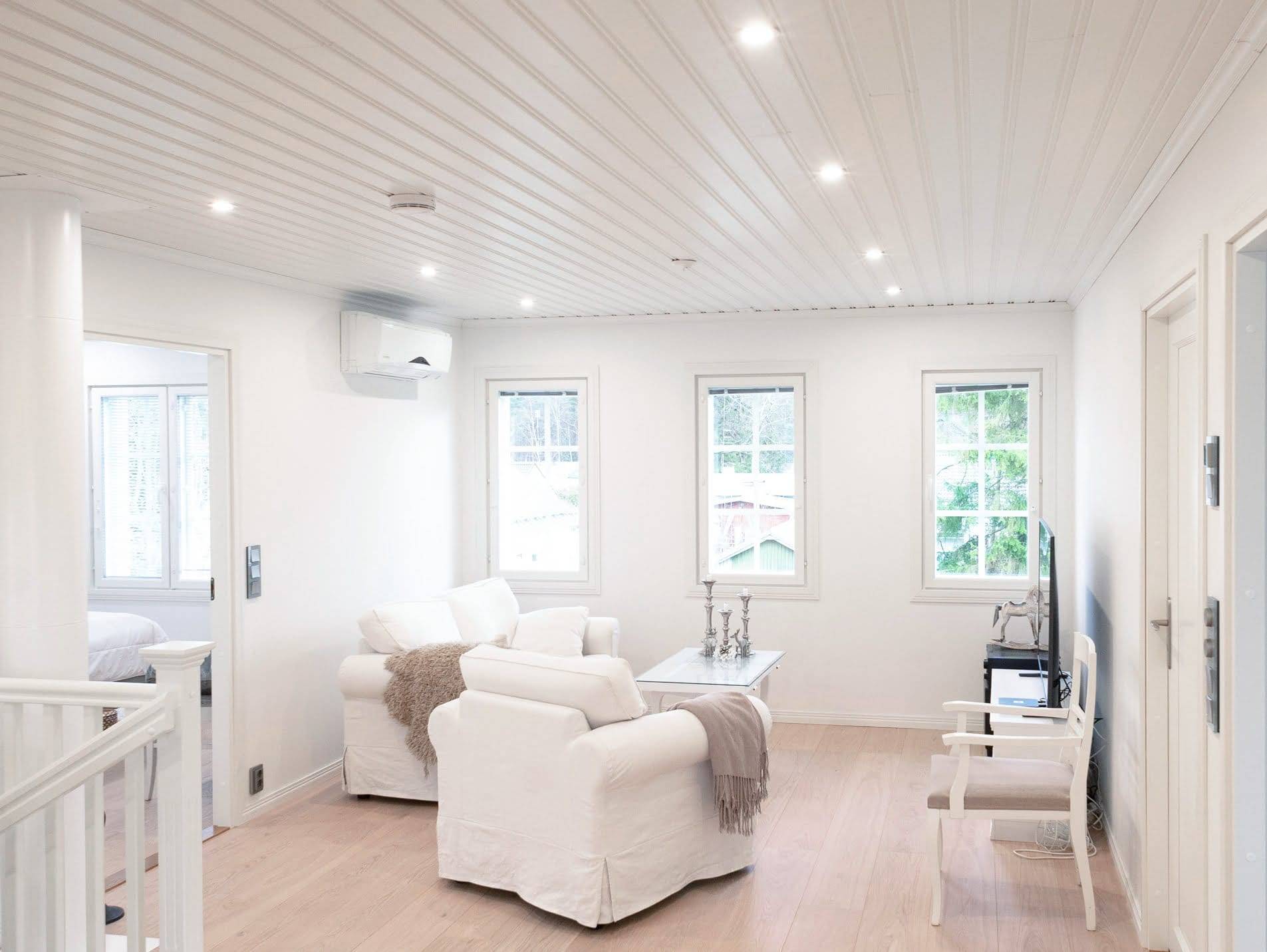 Salon moderne avec éclairage LED, fauteuils blancs, table basse et télévision ; la lumière naturelle provenant des grandes fenêtres accentue le plancher en bois.
