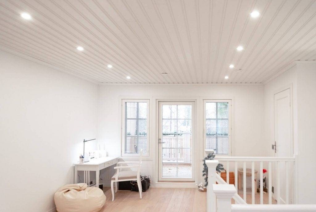 Chambre moderne avec éclairage LED encastré, murs blancs, bureau, chaise, pouf, portes vitrées donnant sur le balcon ; rampe d'escalier à droite.