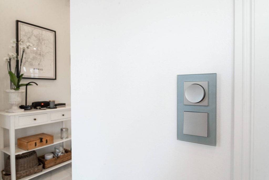 Interrupteur LED moderne avec deux boutons sur un mur blanc ; console blanche avec orchidée en pot et carte géographique encadrée dans une pièce minimaliste.
