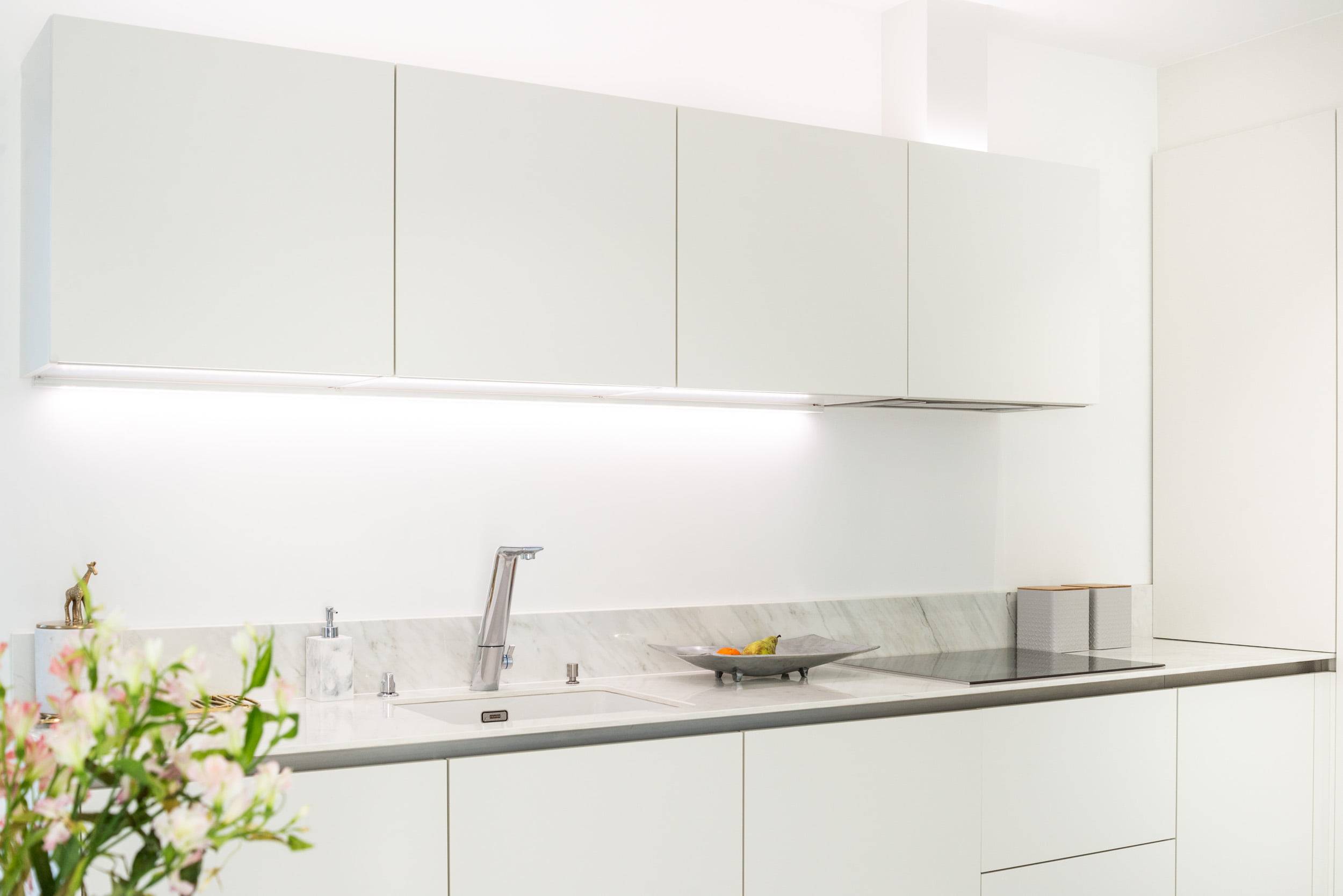 Une cuisine élégante avec des armoires blanches épurées, un plan de travail moderne en marbre et une robinetterie en acier inoxydable. L'éclairage LED sous le comptoir accentue l'aspect de l'espace de travail. Une plante décorative et une coupe de fruits rehaussent l'aspect du plan de travail. Vous pouvez trouver les bonnes solutions d'éclairage LED sur LedStore.fi, où vous trouverez des produits LED de qualité pour compléter l'aspect de votre cuisine.