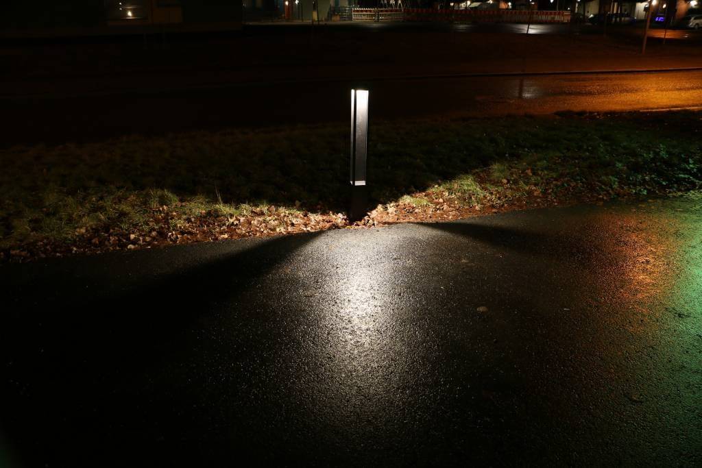Un lampadaire à LED qui éclaire une rue humide avec des motifs triangulaires bordés d'herbe ; une toile de fond urbaine avec des lumières lointaines.
