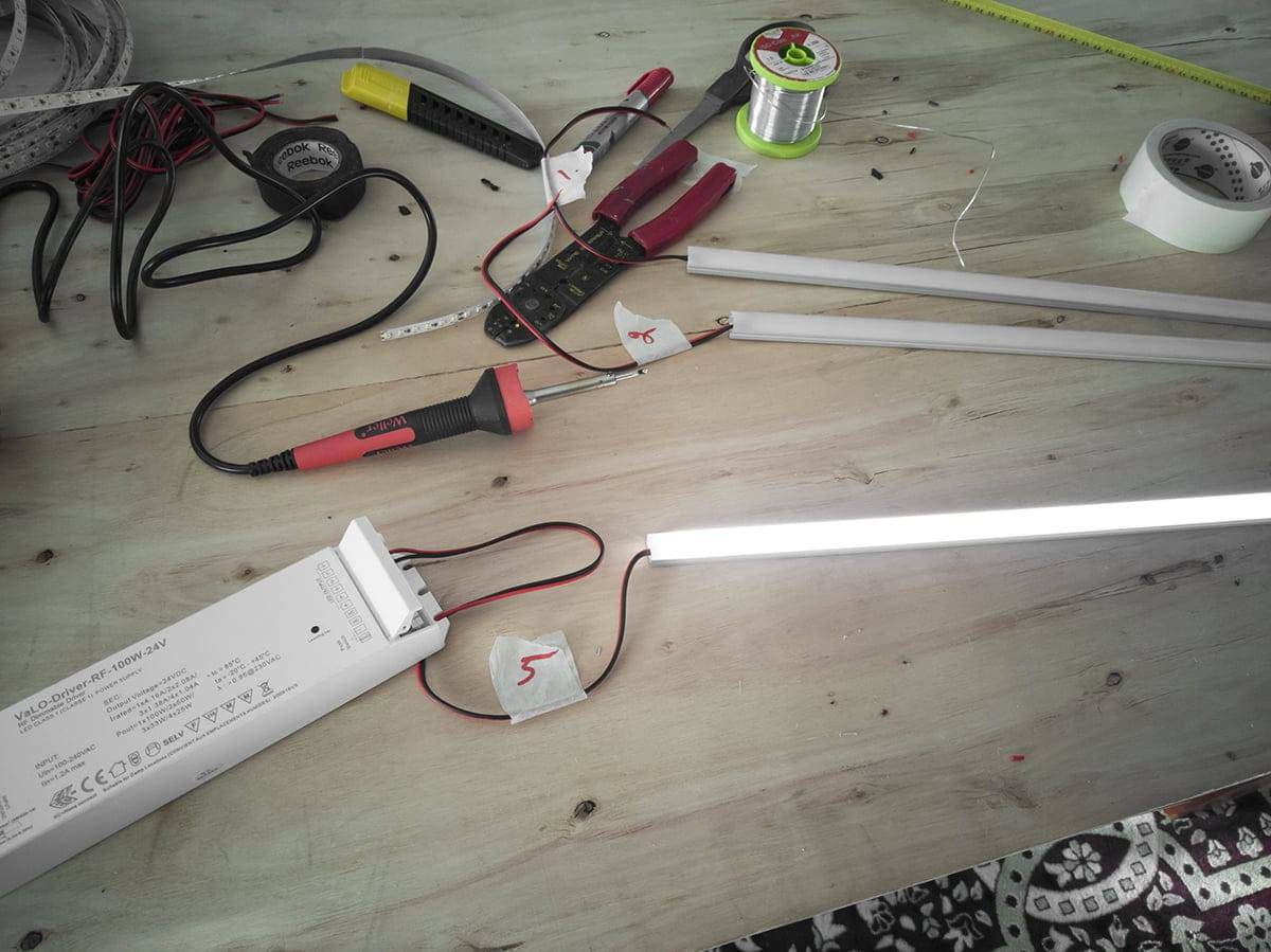 Établi avec des outils électriques, des bandes LED, des fils, des tubes fluorescents lumineux ; comprend des pinces, un multimètre, un fil à souder et une alimentation électrique.