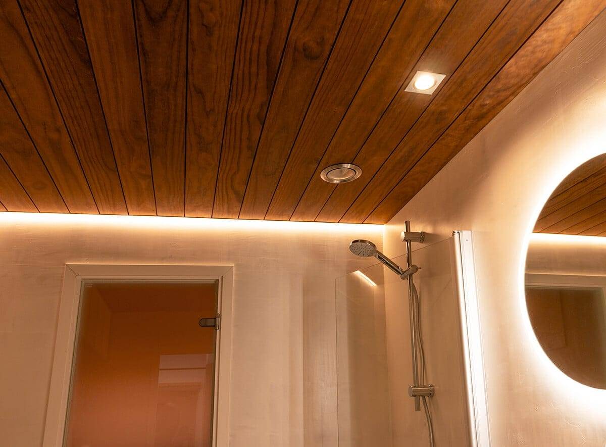 Salle de bain au plafond en bois avec éclairage LED ambiant, pomme de douche moderne, spot lumineux et miroir circulaire LED ; porte à gauche.