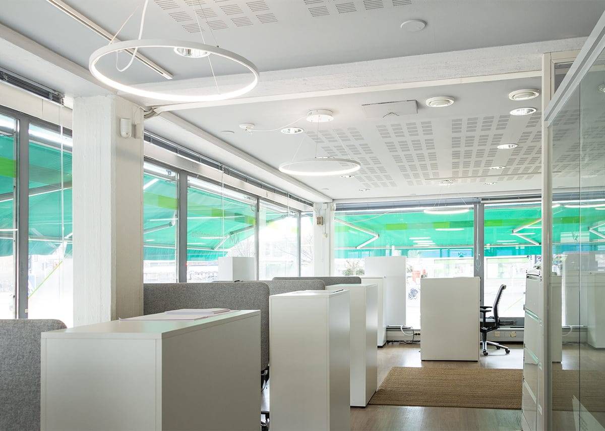 Un bureau moderne avec des bureaux blancs, des chaises grises, des lampes LED rondes et de grandes fenêtres aux teintes vertes. Des sols en bois clair et des tapis agrémentent l'espace.