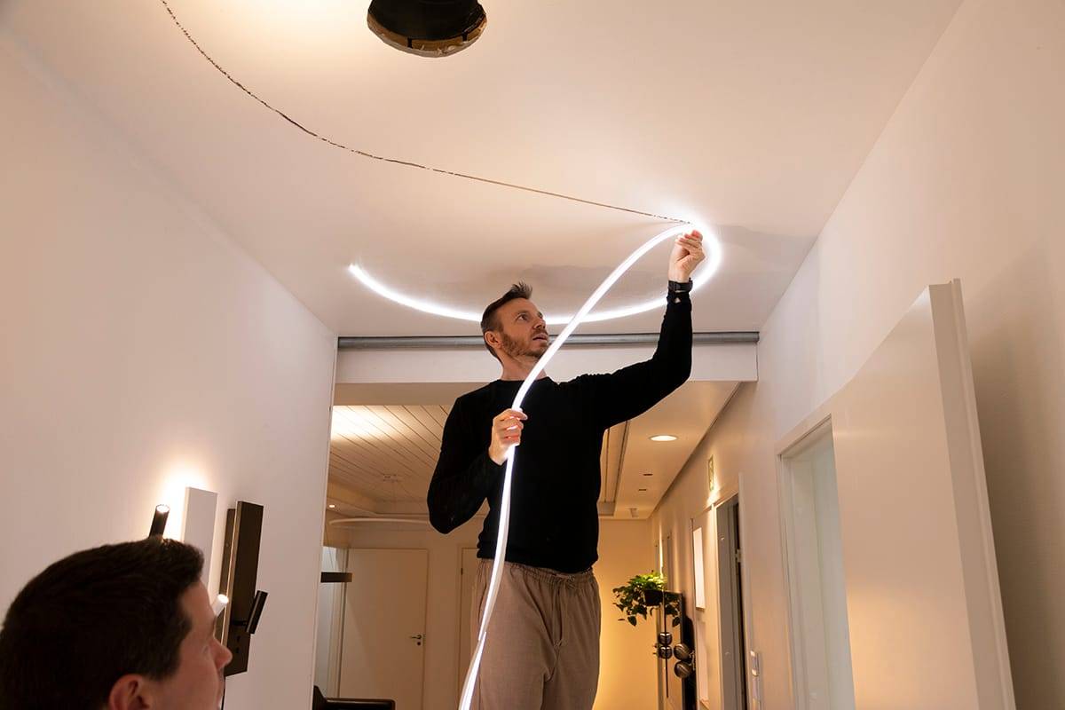 Une personne fixe une bande LED flexible à un plafond moderne, ce qui améliore l'éclairage de la pièce. Pull noir, pantalon beige ; une autre personne au premier plan, floue.