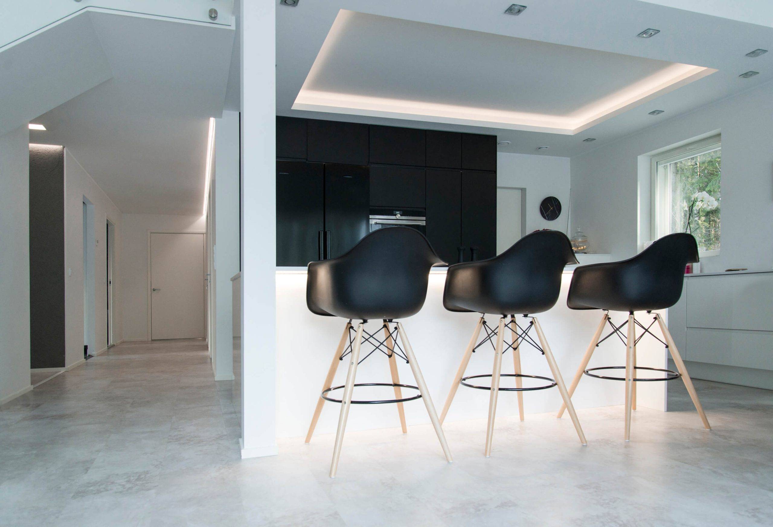 Bande d'aluminium et son utilisation dans l'éclairage LED Une cuisine moderne avec des tabourets de bar noirs sur un îlot blanc, des armoires sombres, un plafond encastré avec éclairage LED, une fenêtre et un couloir minimaliste créent un look élégant.