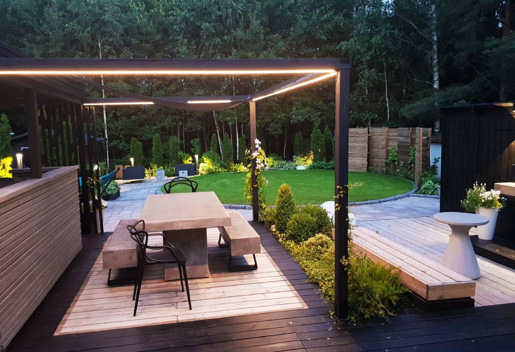 Un patio moderne avec une table à manger en béton et des chaises noires sous une pergola minimaliste. La pergola est dotée d'un éclairage LED intégré qui crée une atmosphère sombre. L'espace est entouré d'une végétation verte, d'une pelouse unique et d'un chemin en pierre, ce qui ajoute au charme naturel. En outre, comme petit détail, il y a une jardinière moderne blanche pour profiter de l'élégante terrasse. Découvrez la vaste gamme d'éclairages LED de LedStore.fi et apportez la même élégance moderne à votre propre jardin !