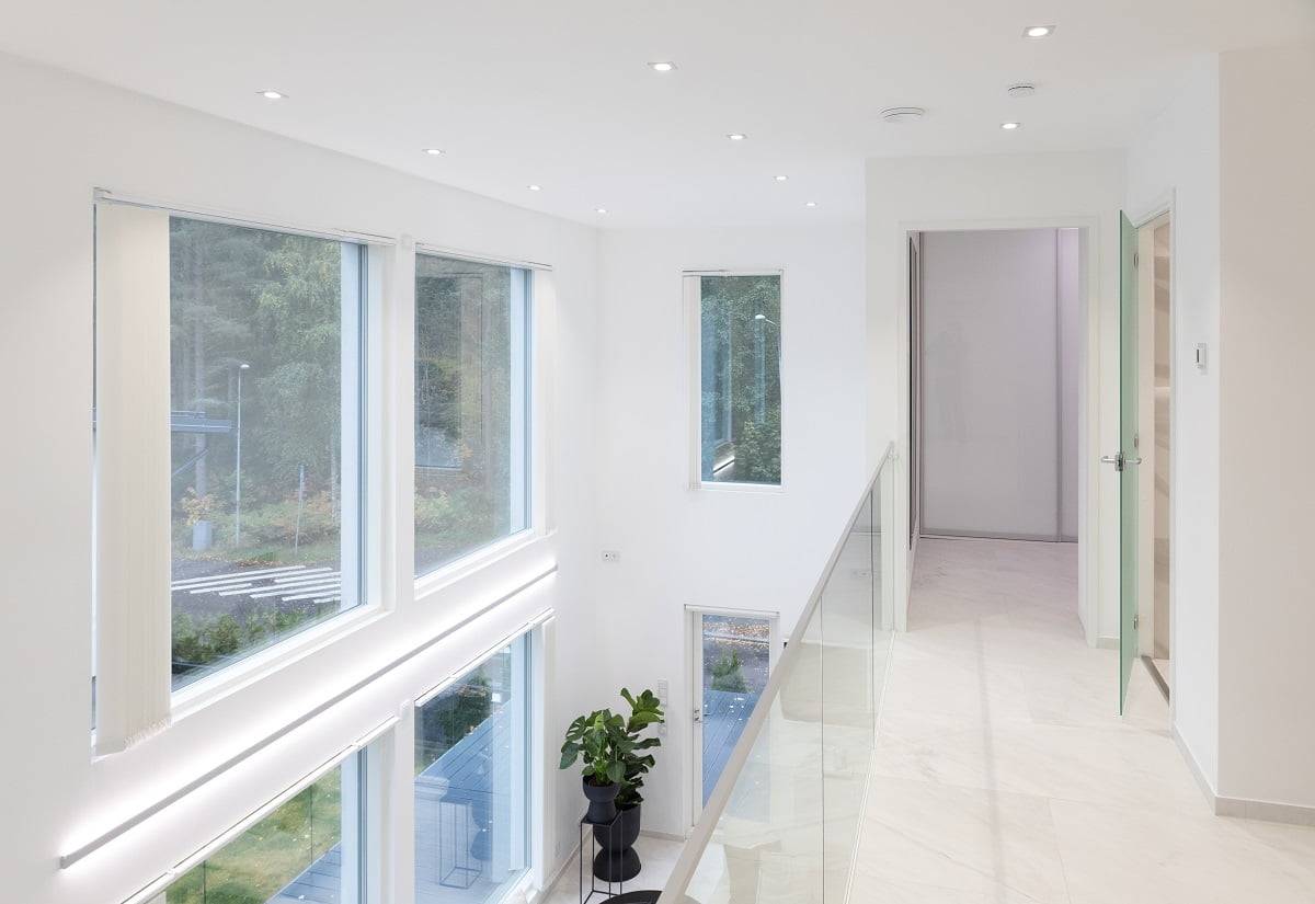 Un couloir intérieur lumineux et moderne avec de grandes fenêtres des deux côtés permet à la lumière naturelle de pénétrer à l'intérieur. Les murs blancs et l'éclairage LED encastré créent une atmosphère conviviale pour les enfants. Une balustrade en verre ajoute de l'élégance. Une plante verte en pot apporte une touche de couleur à l'ensemble. Bienvenue dans la gamme d'éclairage LED de haute qualité de LedStore.fi et créez l'espace lumineux de vos rêves !
