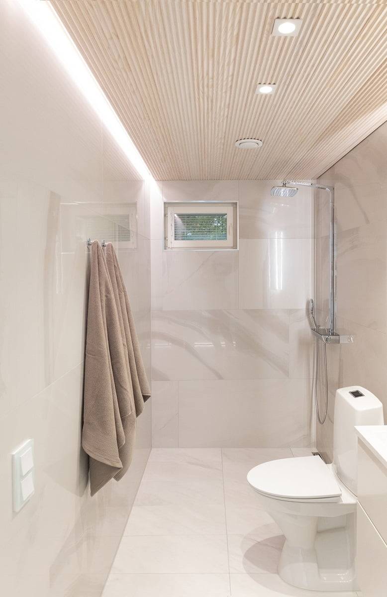 Une salle de bains moderne et minimaliste avec des murs beige clair et un plafond en carreaux de bois. Un porte-serviettes mural avec deux serviettes, une petite fenêtre et une douche séparée par une cloison en verre ajoutent de l'élégance à l'espace. Des plafonniers LED intégrés assurent un éclairage efficace. LedStore.fi : Découvrez les solutions d'éclairage LED qui conviennent parfaitement aux intérieurs élégants et intemporels.   