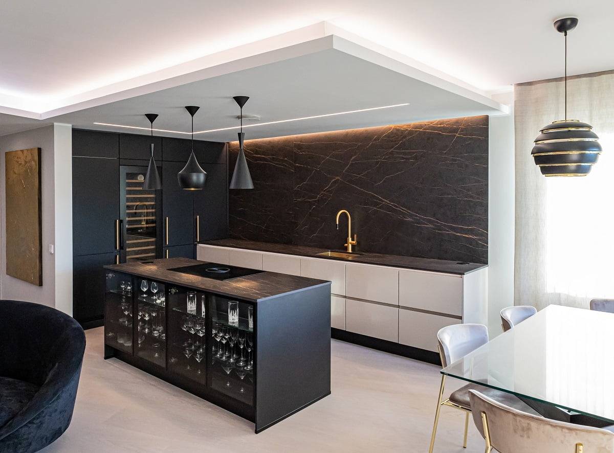 Une cuisine moderne avec des carreaux noirs et blancs, des carreaux blancs, un plan de travail en marbre noir et un robinet doré. Des lampes suspendues éclairent un îlot noir avec de la verrerie qui coule. Une table de salle à manger adjacente en verre avec des chaises rembourrées est surmontée d'une suspension noire et dorée éclairée par des LED. Découvrez la vaste gamme d'éclairages LED de LedStore.fi pour créer un look similaire dans votre propre maison !