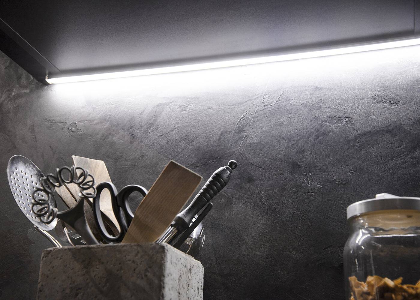 Photo de cuisine avec de la vaisselle sur un support, une bande lumineuse LED met en valeur le mur sombre et texturé ; un bocal en verre est visible sur la droite. Idéal pour un éclairage LED moderne.