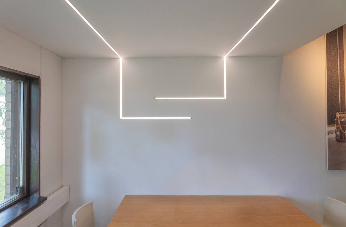 Une pièce minimaliste avec une table et une chaise en bois reçoit un nouveau look grâce aux bandes LED élégantes de LedStore.fi, qui forment des motifs horizontaux et verticaux sur le mur. La lumière de la fenêtre à gauche complète l'éclairage, créant un aspect géométrique moderne contre les notes en arrière-plan.