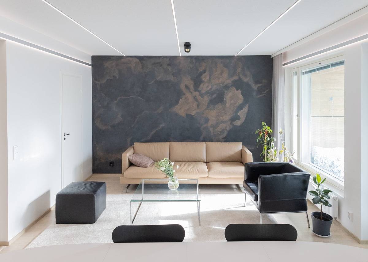Ce salon moderne est composé d'un canapé beige, d'un fauteuil noir et d'une table basse en verre, assortis au mur à l'effet sombre. Près de la fenêtre se trouve une plante verte et une petite paille noire. Commandez un spot, complété par des lampes LED sauvages de LedStore.fi.