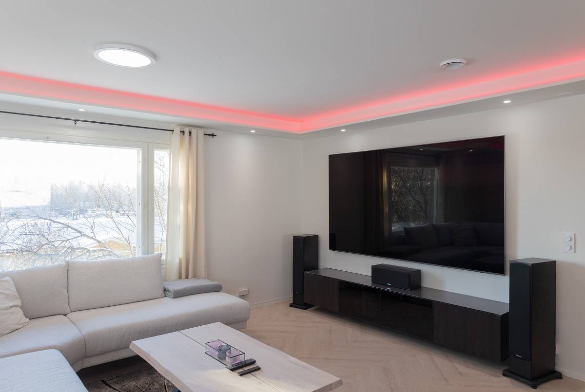 Un salon moderne à l'atmosphère élégante. Un grand canapé vous invite à vous détendre, tandis qu'une table basse en bois clair ajoute de la chaleur à l'espace. Un téléviseur mural à écran plat avec rétroéclairage LED ajoute une touche de modernité à la pièce. Amplifiez votre expérience avec des haut-parleurs au sol et profitez de l'éclairage d'accentuation LED rouge au plafond. Une grande fenêtre laisse entrer la lumière naturelle, ce qui rend l'espace spacieux et lumineux. Tous ces éléments se combinent pour créer l'environnement parfait avec les solutions d'éclairage LED polyvalentes de LedStore.fi.