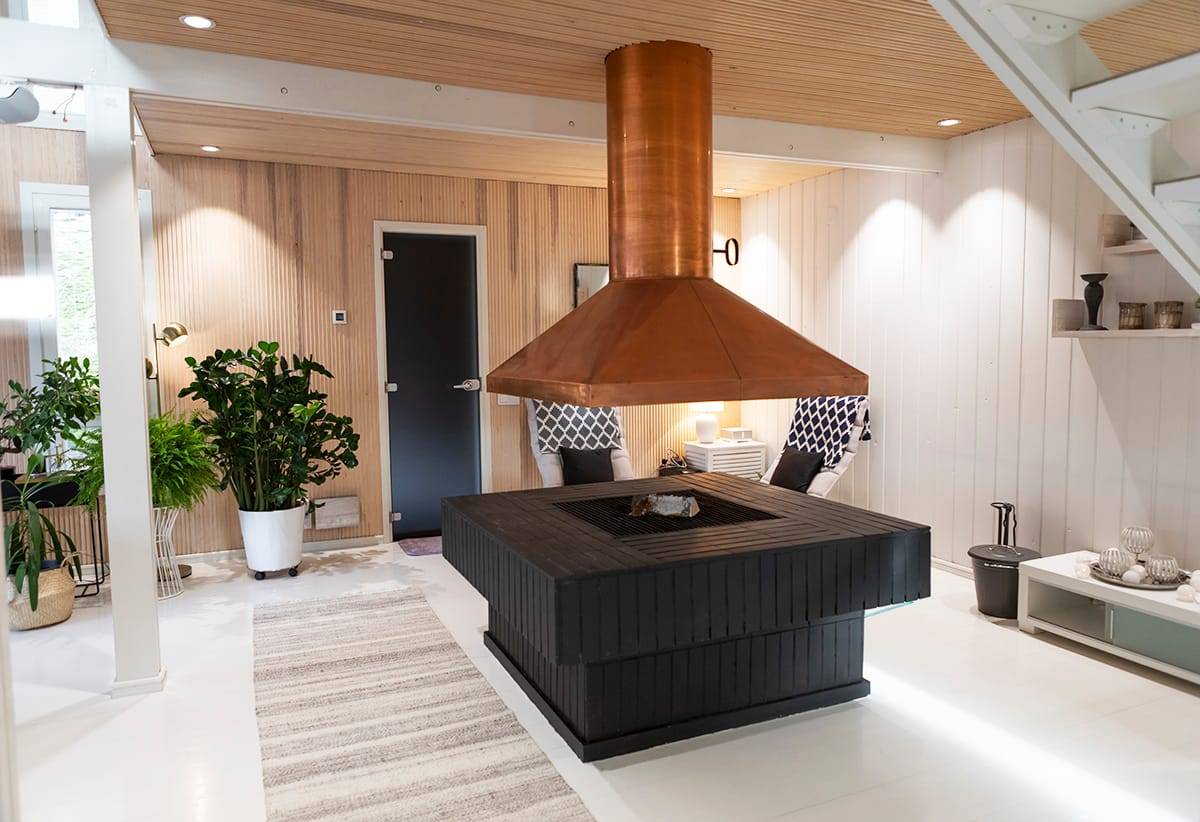 Salon moderne avec cheminée noire et bois au centre, tuyau en cuivre, éclairage LED, fauteuils rembourrés, verdure et décoration élégante.