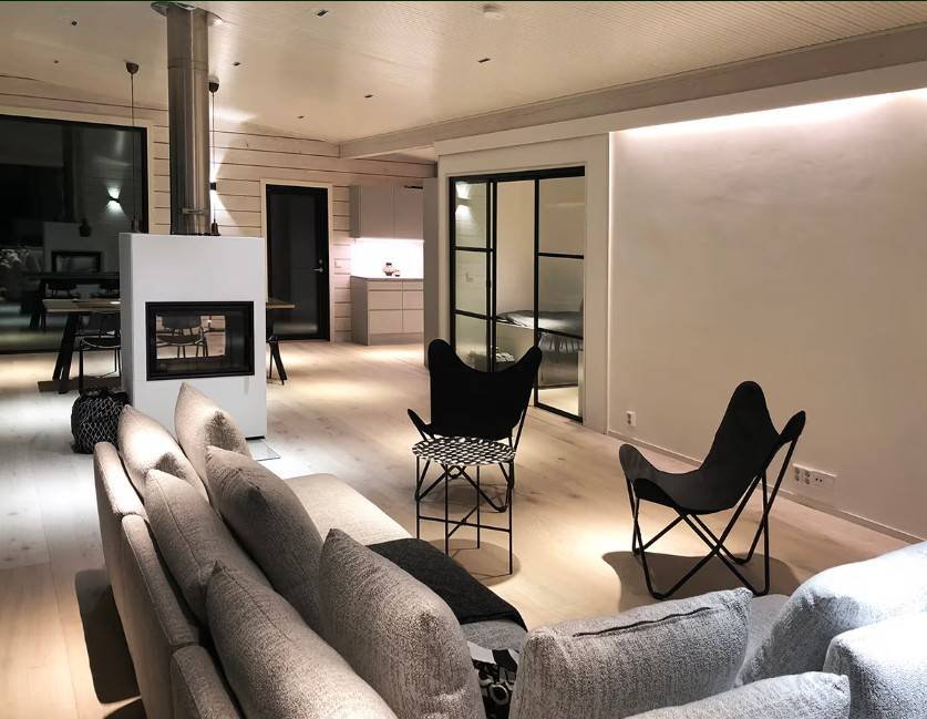 Angle d'incidence de la lumière - effet sur l'éclairage Un salon moderne avec un canapé gris, des chaises noires, une cheminée et une cuisine ouverte en arrière-plan - l'atmosphère et le style sont créés par la solution d'éclairage LED de haute qualité de LedStore.fi.