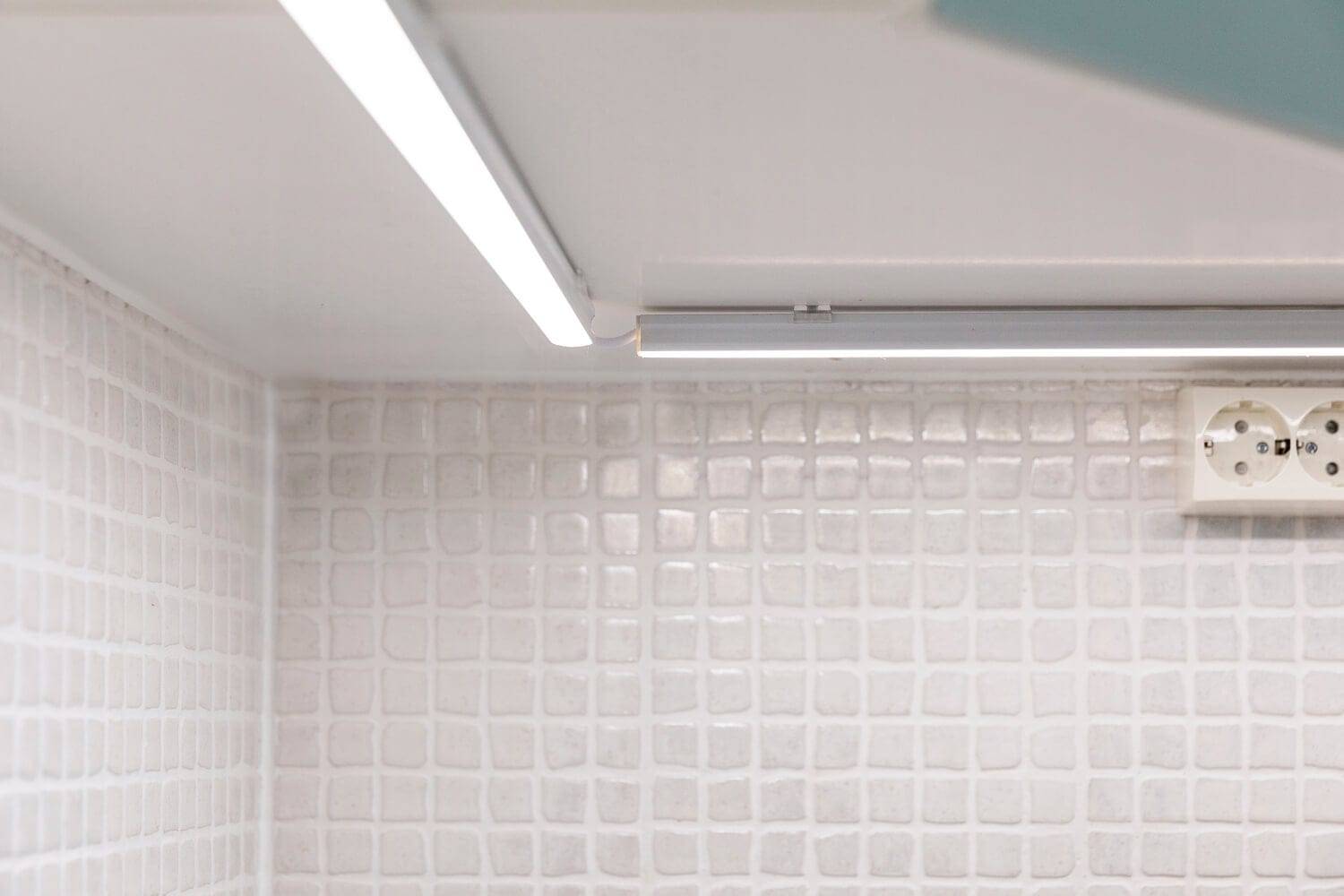Une crédence de cuisine moderne composée de carreaux blancs texturés, éclairée par un éclairage LED linéaire placé sous le meuble ; une douce lueur met en valeur la prise murale.