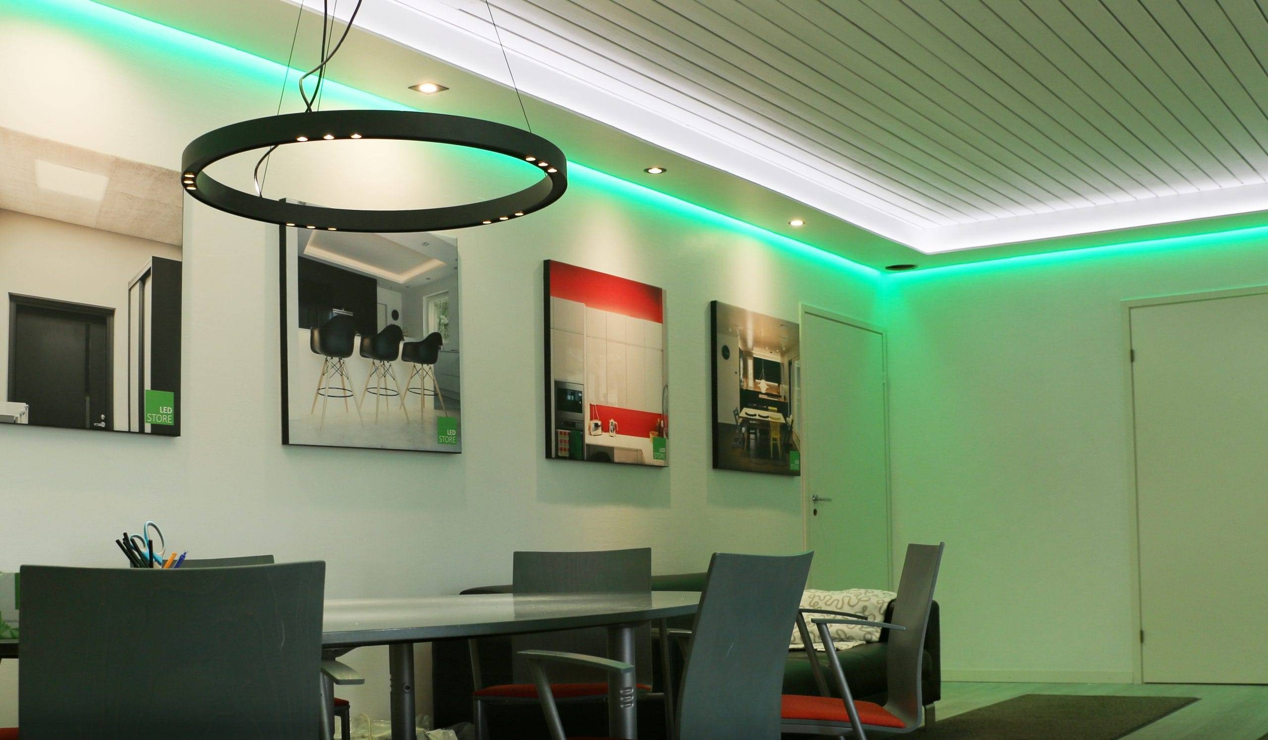 Contrôle de l'éclairage Zigbee partie 2 - Revue des dispositifs Zigbee et de l'appairage Une pièce moderne avec une lampe LED circulaire noire, des chaises grises autour d'une longue table, des œuvres d'art encadrées au mur et un éclairage au plafond avec des reflets verts pour créer une atmosphère élégante.
