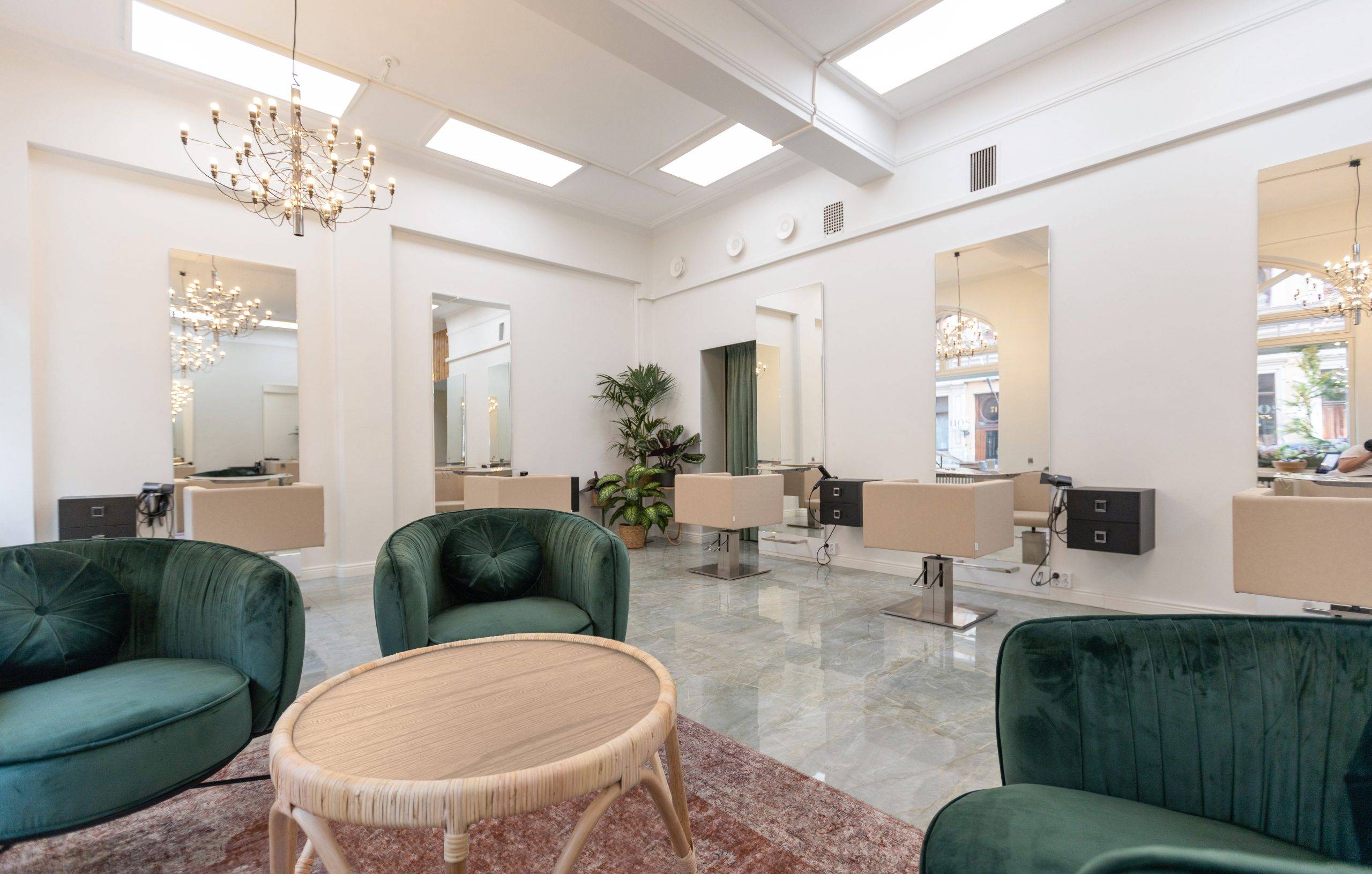Un intérieur de salon moderne et lumineux avec des chaises en velours vert entourant une table ronde avec un tapis rouge. Des postes de travail avec des miroirs et des chaises beiges sont alignés le long des murs. Ils sont éclairés par des lustres LED fixés au plafond. De grandes fenêtres laissent entrer la lumière naturelle, tandis que la verdure renforce l'ambiance de l'espace. Dans ce type d'environnement, les solutions d'éclairage LED de haute qualité de LedStore.fi sont idéales, créant l'équilibre parfait entre la lumière et les économies d'énergie sans compromettre l'élégance.