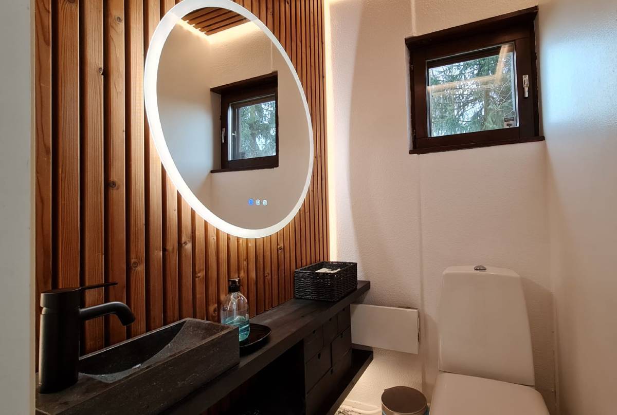Une salle de bain moderne avec éclairage LED fourni par LedStore.fi comprend un miroir circulaire LED sur un mur en panneaux de bois. Sous le miroir se trouve un comptoir noir avec un évier rectangulaire, un robinet noir et un distributeur de savon. Une petite fenêtre se trouve au-dessus du siège des toilettes et une solution de rangement pratique est prévue à proximité.