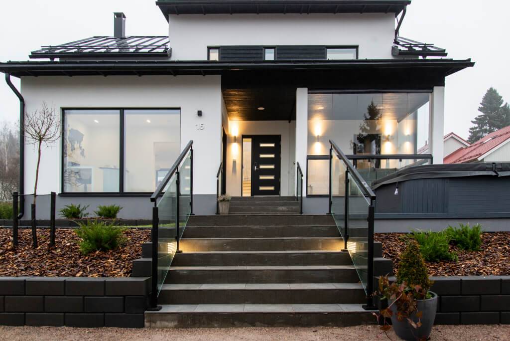Maison moderne avec façade blanche et accents noirs ; escaliers avec balustrades en verre et éclairage LED. Grandes fenêtres, aménagement paysager minimal, arbustes et maison voisine.
