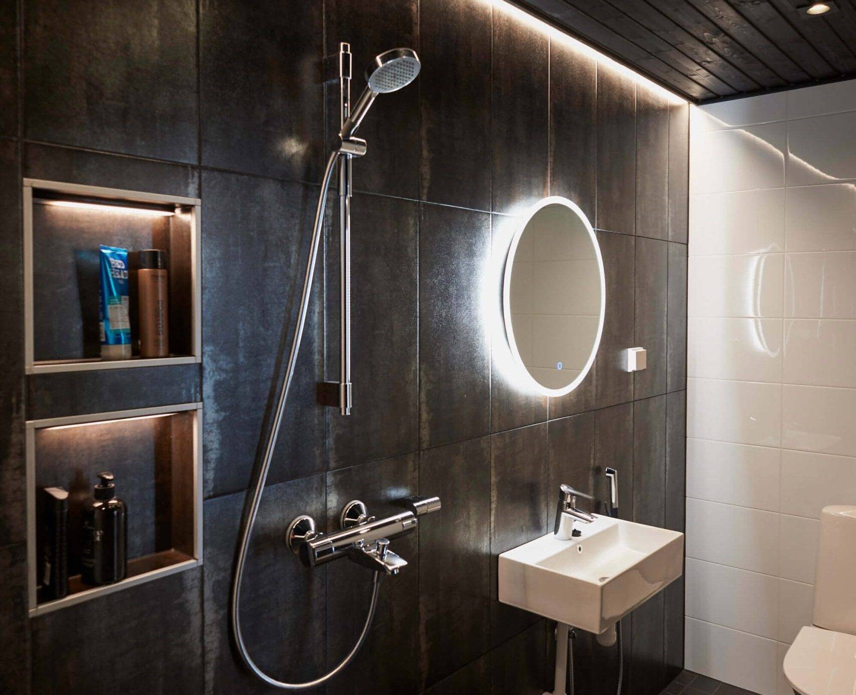 Salle de bains élégante avec carrelage foncé, miroir rétroéclairé par LED au-dessus du lavabo, douche, étagères encastrées et plafond noir. Des toilettes modernes complètent l'ensemble.