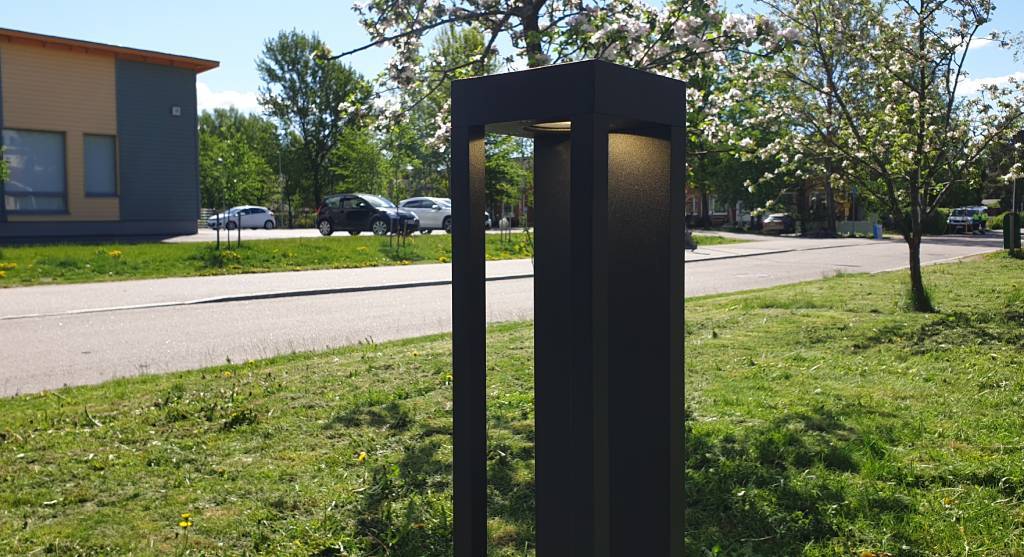 Un lampadaire noir moderne avec une lumière LED le long de la route ; des arbres en fleurs, des voitures en stationnement et un bâtiment avec de grandes fenêtres en arrière-plan sous un ciel ensoleillé.
