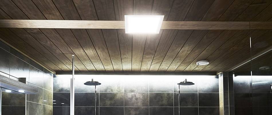 Salle de bains moderne avec plafond en bois, luminaire LED carré, murs carrelés foncés, deux pommeaux de douche à effet pluie et une atmosphère chaleureuse grâce à un éclairage LED doux.